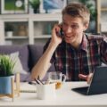 Man in a checkered shirt talks on phone in home office setting with laptop and coffee.
